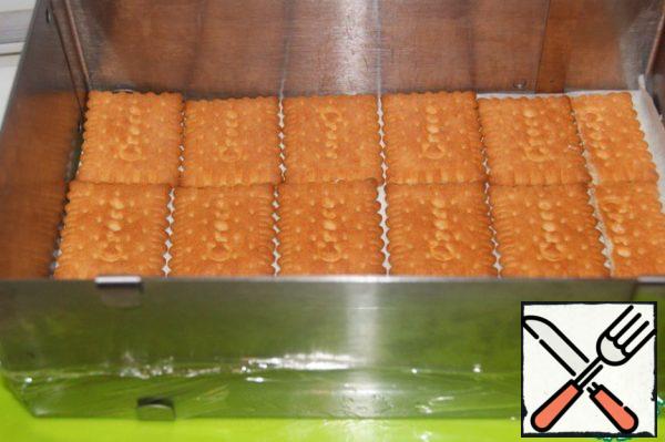 Prepare a detachable form of 26x22 cm (I have a frame) and, so that there are no leaks, I wrapped the frame with cling film. Cover the bottom with baking paper and lay out a row of cookies.
