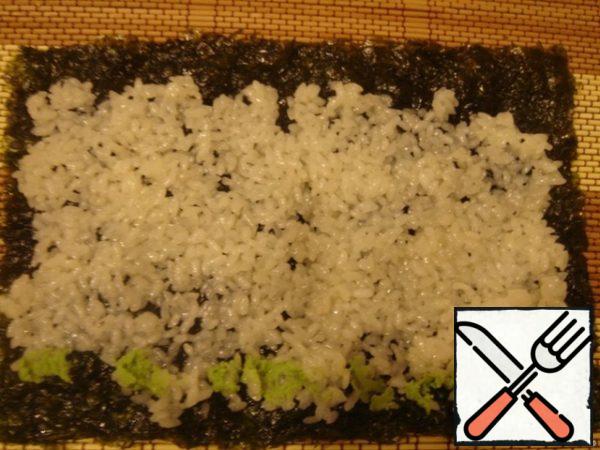 Now you can start cooking rolls.
On the Mat for sushi (makisu) put a plastic wrap on top of a sheet of nori.
On it lay a thin layer of rice. To make it easier to do, hands need to be moistened with water.
Over rice in the center or from one edge it is necessary to put a thin strip of wasabi. Put the filling-cucumber, cheese, salmon.