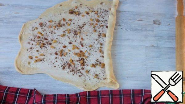 The first piece of dough is rolled out into a thin long layer. It can be stretched, it stretches well. Spread softened butter over the dough, sprinkle with cinnamon and raisins. Roll the dough into a tight roll. Put the first roll under the towel. And we do the same with the second piece of dough.

