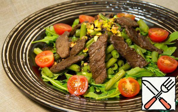 We assemble the salad: lay out a pillow of arugula, put a mixture of beans and lentils on it, garnish with pieces of beef, pistachios and cherry tomatoes.
