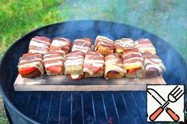 Wrap the slices of bread with strips of bacon filling.
Once the temperature of the coals has reached optimum, place the grill Board on the grill grate, over the coals, face down. In this position, hold the Board for 1-2 minutes until the smoke goes (the dried front side will not allow the grill Board to bend excessively during baking).
Then turn the grill Board face up and place the bacon rolls on top.