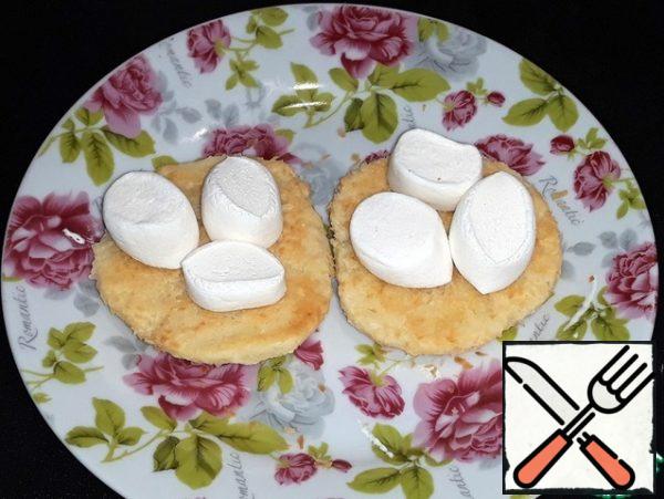 At the bottom of the cookie, put the marshmallows cut into three parts. In the microwave oven, we heat it up for a few seconds until the marshmallows increase in size.
