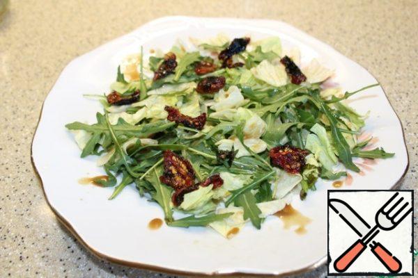 On the dish, put the large torn iceberg lettuce leaves and arugula leaves, dried tomatoes, sprinkle with a small amount of dressing.