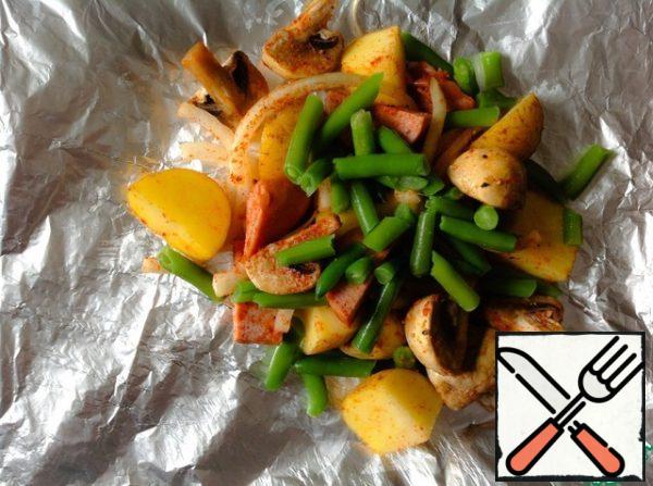 Cut out four sheets of foil approximately 30x30 cm in size. If the foil is thin, form a square in two layers.
Divide the sausages, potatoes, green beans, mushrooms and onions into 4 equal parts and place in the center of each square of foil in a single layer.