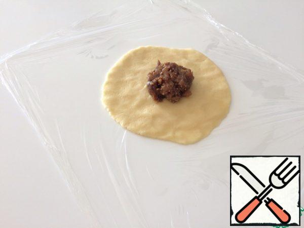 We tear a square piece from the roll of food film. We spread a ball of dough on it and press it with our hands into a flat cake about 8 cm in diameter. Top with a teaspoon of filling.