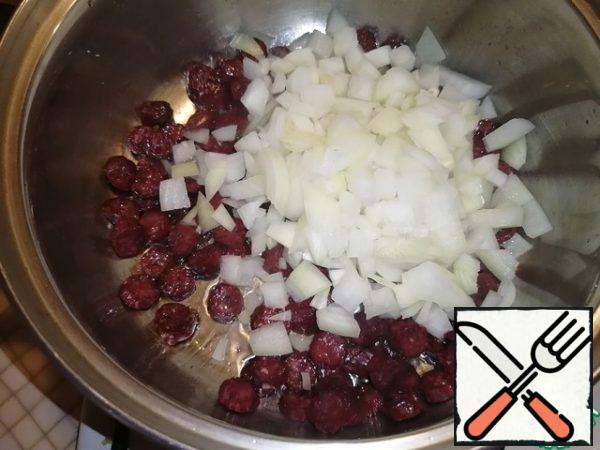 Cut the onion into cubes. Pour out to the sausages. Fry for 4-5 minutes.