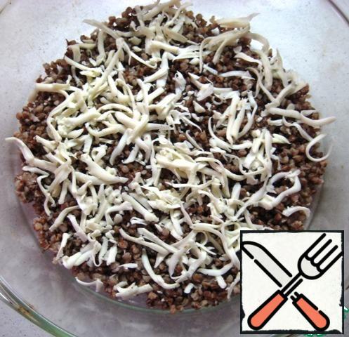 In a deep baking dish, put half a portion of buckwheat, flatten, and distribute 1/3 of grated cheese on top.