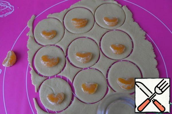 We take out the dough, roll it into a layer 5 mm thick, cut out the circles. I used a regular glass. The diameter of the circle should be larger than the tangerine. On a circle of dough, put a slice of tangerine on the edge.