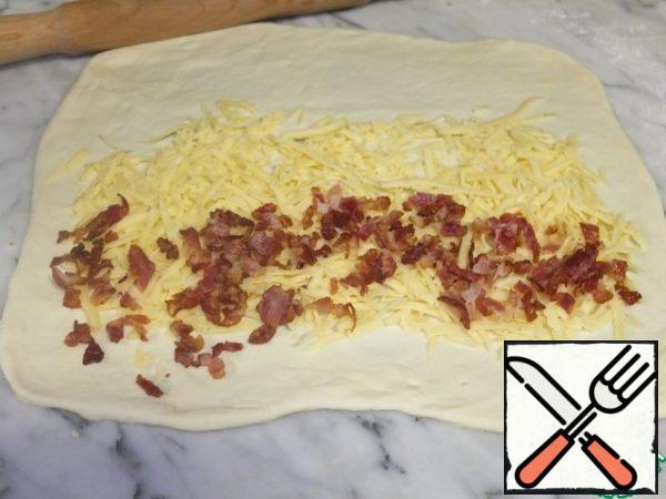 
After an hour, knead the dough and divide it into two or three parts, as you prefer.
I divided the dough into three parts to make it easier to roll out.
The dough is elastic, rolls out well, does not stick.
Roll out each part into a rectangle.
Lay out some of the cheese and some of the bacon.
We roll it into a tight roll.