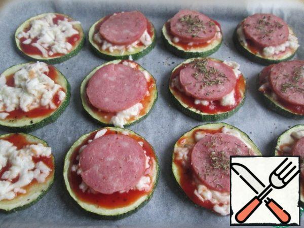 Remove the baking sheet and lightly brush each piece of zucchini with ketchup (or tomato sauce). I used to cook with ketchup for barbecue.
Sprinkle with cheese.
Place a thin circle of sausage on top and sprinkle with spices to taste: oregano, thyme, basil or a mixture of them. I have thyme and basil here.
Put the baking sheet back in the oven for another 5 minutes.
