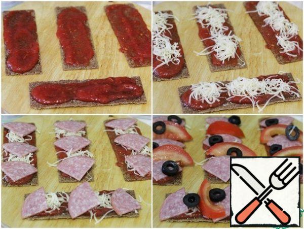 Grease the loaves with the sauce, about 1 tablespoon per loaf.
Sprinkle a small amount of grated cheese on top.
Lay out the sausage slices, add the olives and tomatoes.